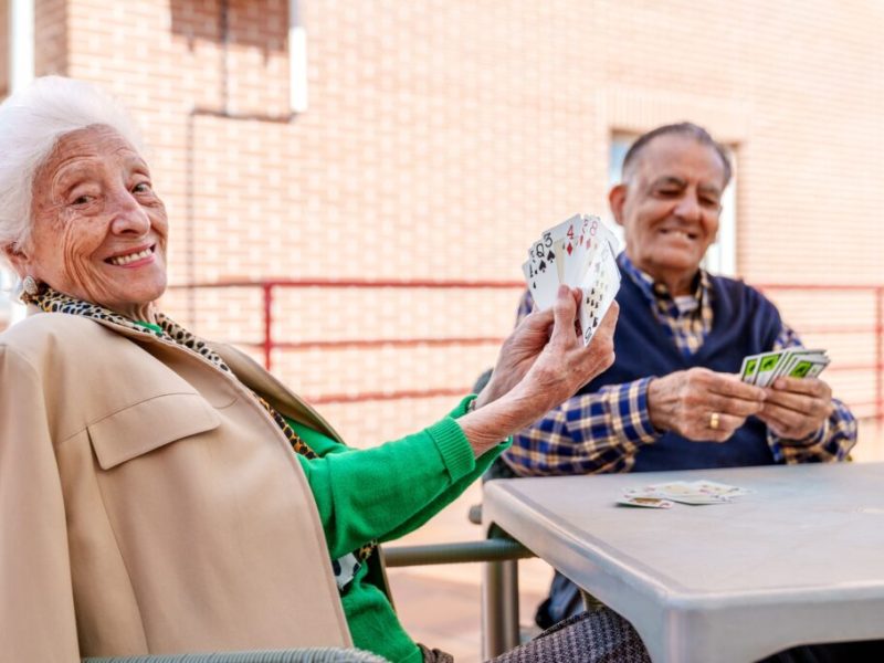In the sunny geriatric patio, a cheerful elderly couple engage in a card game, exuding joy and contentment amidst the vibrant ambiance.