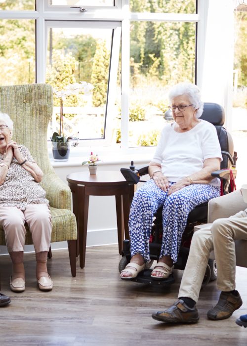 Male And Female Residents Sitting In Chairs And Talking In Lounge Of Retirement Home